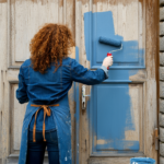 A person painting an interior door with a roller, with a paint can and brush nearby.